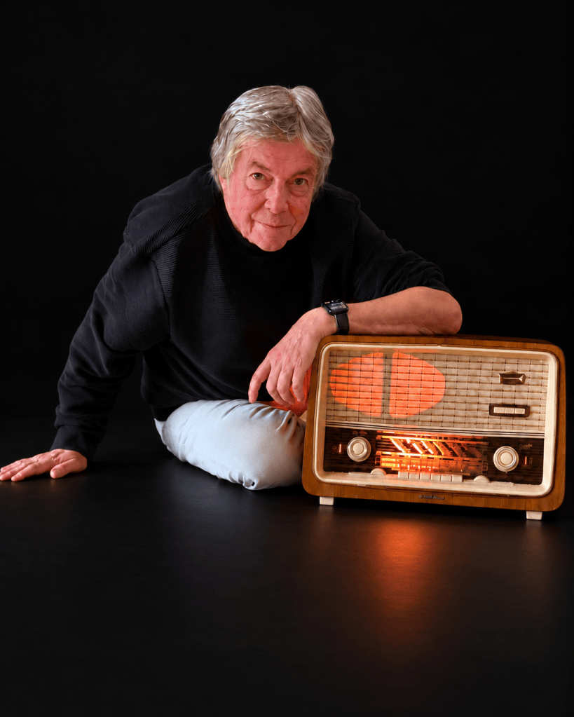 Daniel Duret assis près d’une radio TSF éclairée, sur fond noir.